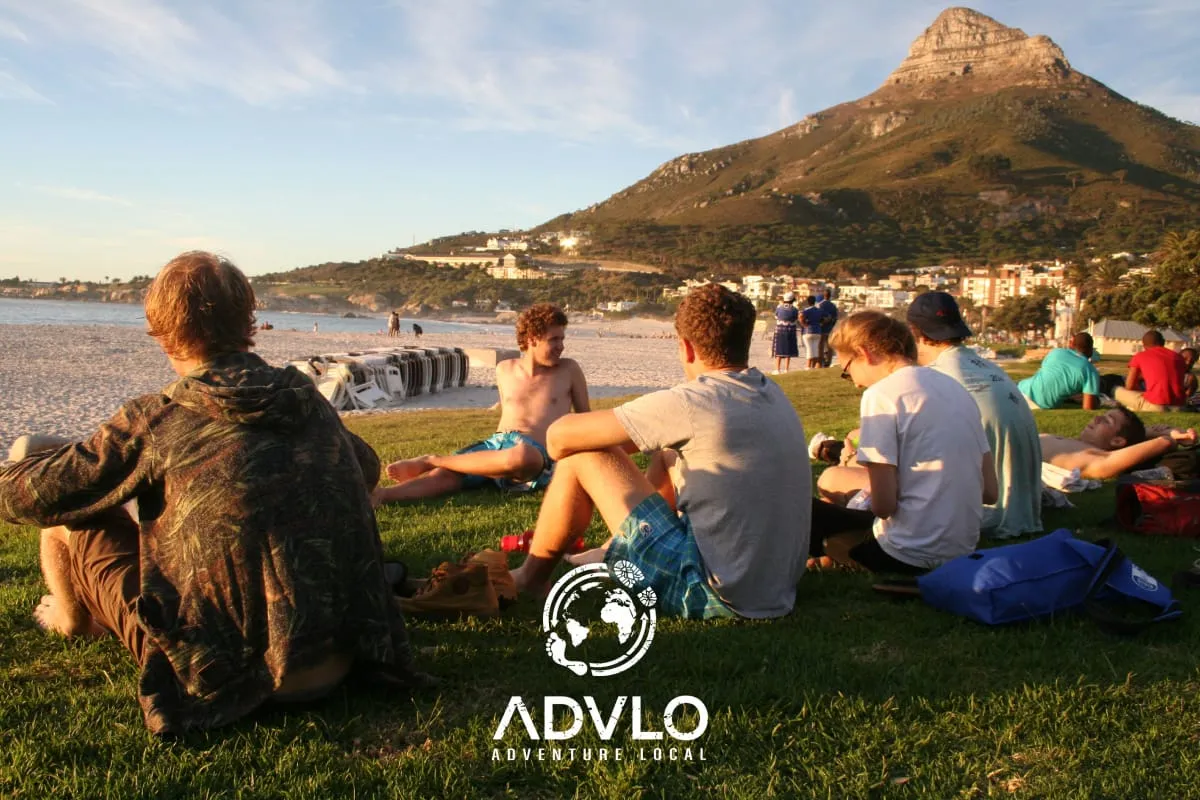 Travelers sitting on a grassy beach below a mountain, with the Advlo logo
