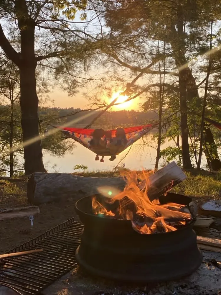 Hammock between trees above a campfire at sunset by a lake