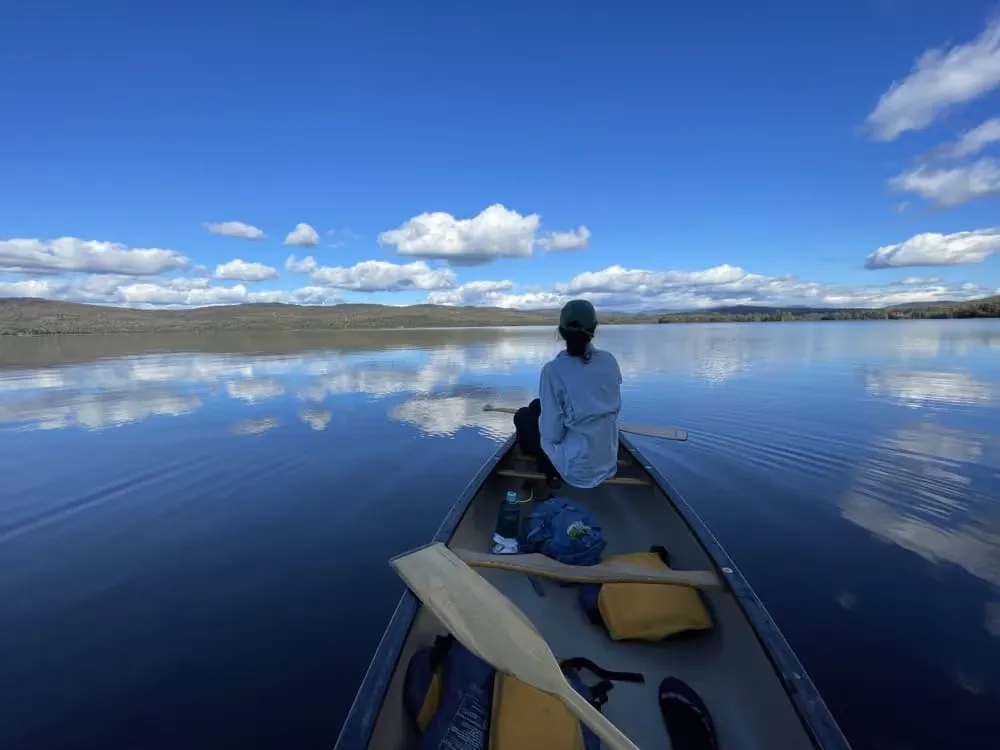View from the back of a canoe toward a paddler on a glassy lake reflecting clouds