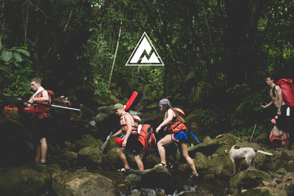 Group hiking through a jungle carrying rafting gear, with the Made by Adventure logo